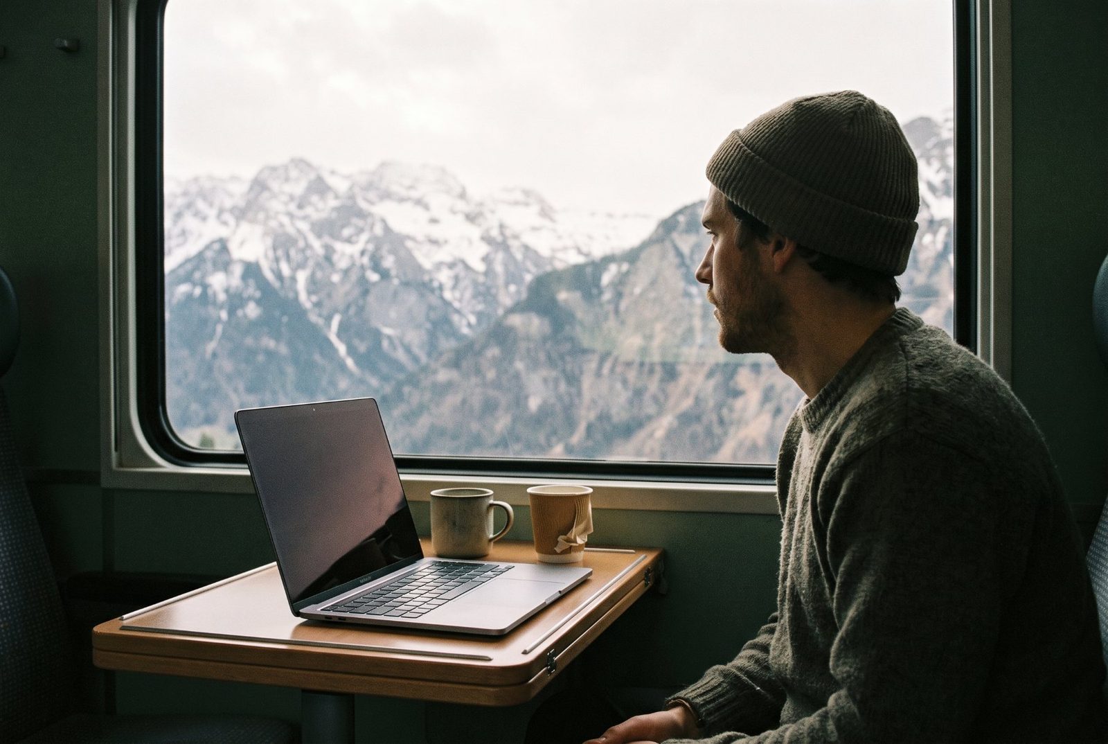 Ein Mann im Zug schaut hinaus auf verschneite Berge, sein MacBook liegt offen vor ihm auf dem Tisch.